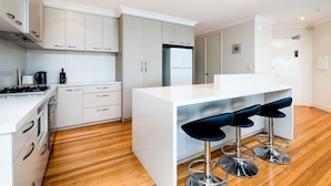 A modern kitchen with white cabinetry, a central island with bar stools, a built-in oven, and a refrigerator. - Fremantle Harbourside  Apartments (Fremantle)