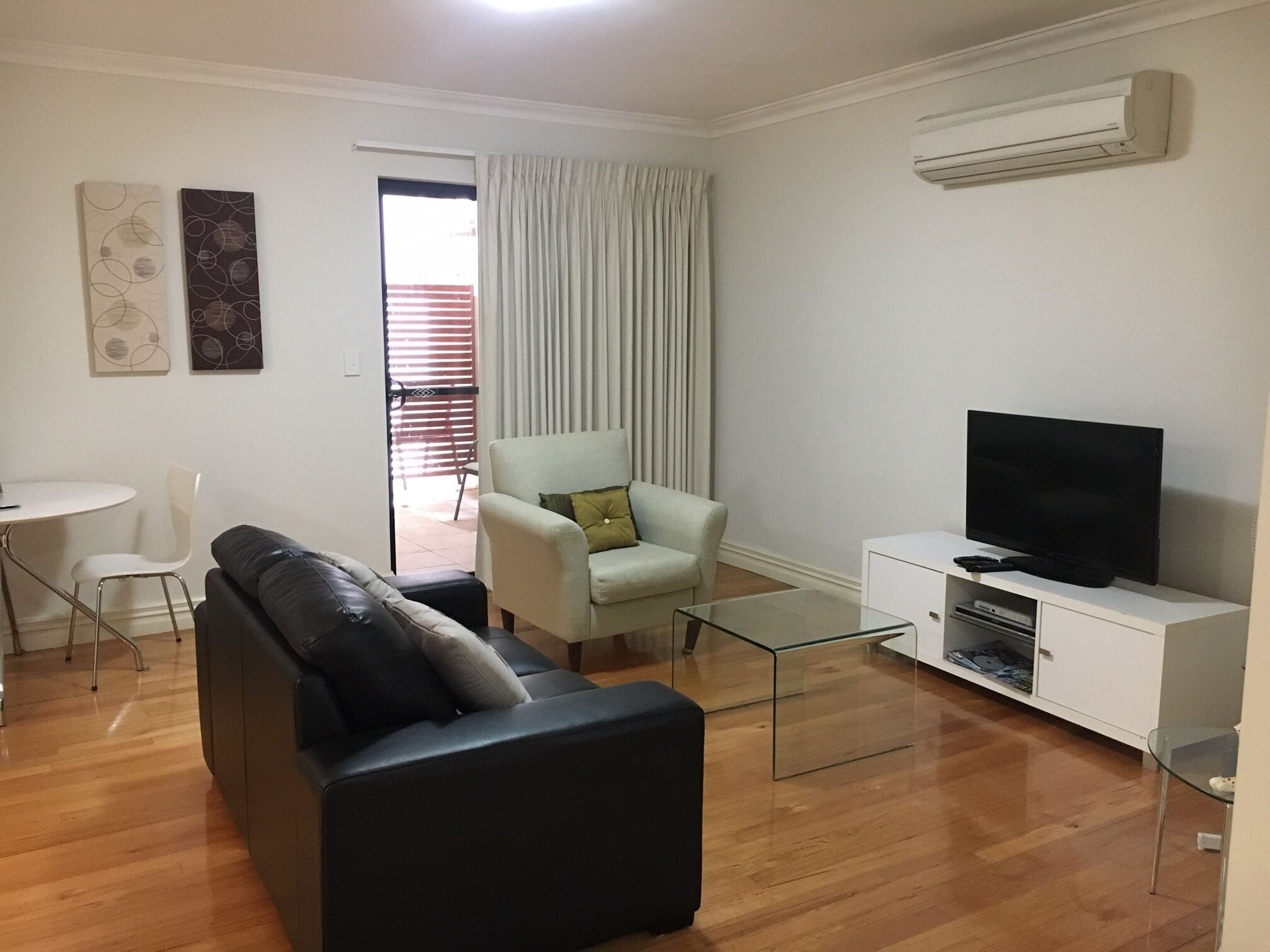 A modern living room with a black leather sofa, a light-colored armchair, a glass coffee table, a flat-screen TV on a white cabinet, and an air conditioning unit on the wall.