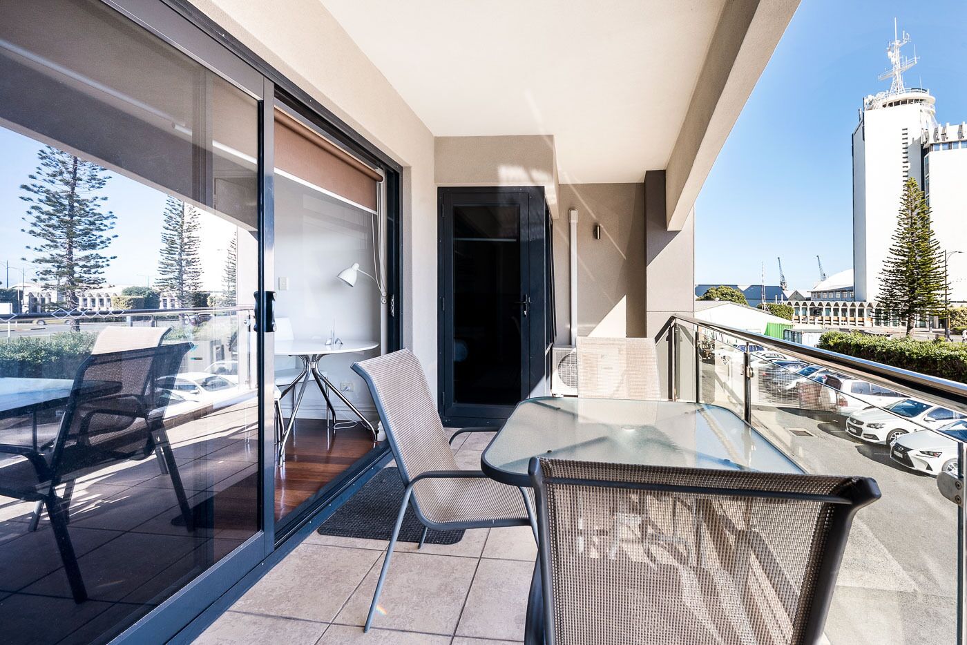 A balcony with a glass table and chairs, a sliding door, and a view of a cityscape including a tall building.