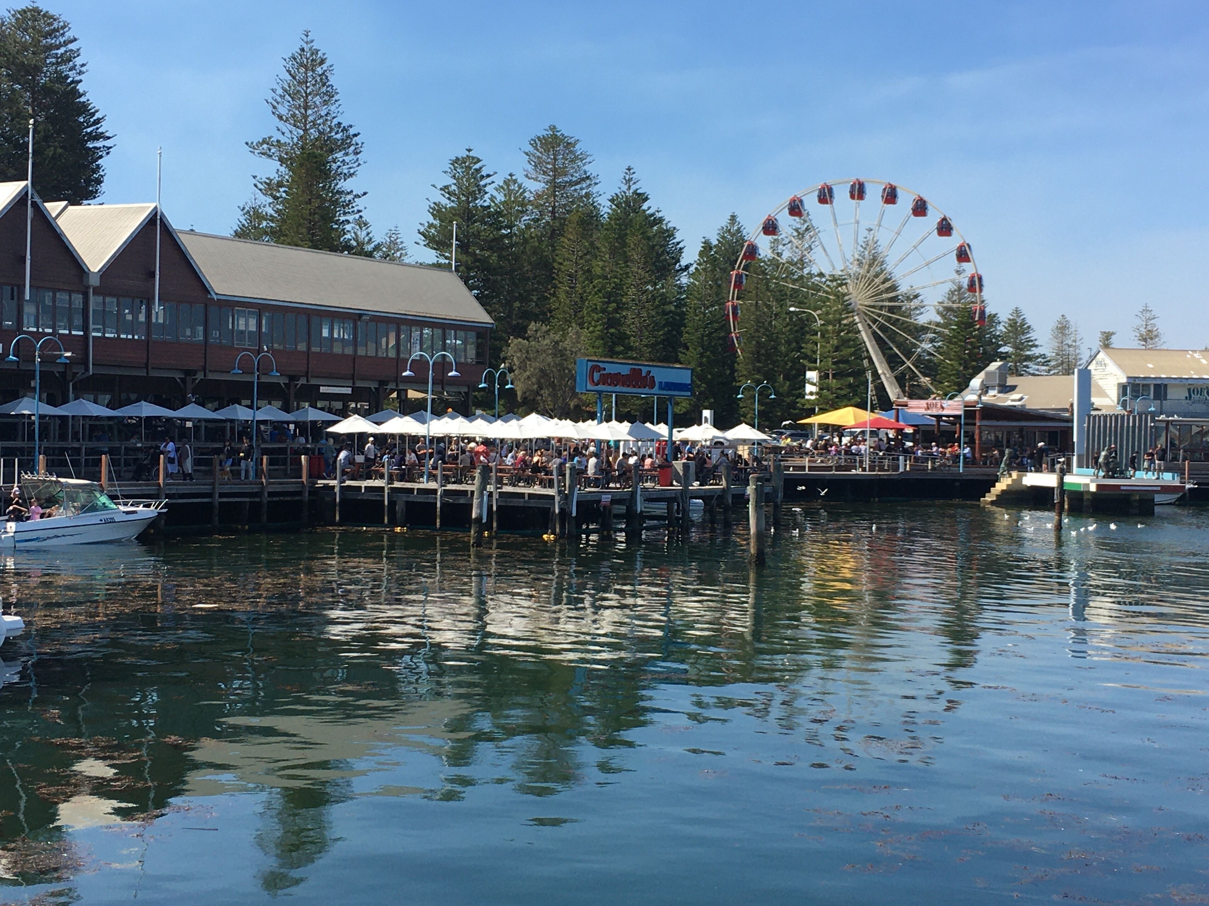 A waterfront area with a Ferris wheel, a dock with people, and a building with outdoor seating.