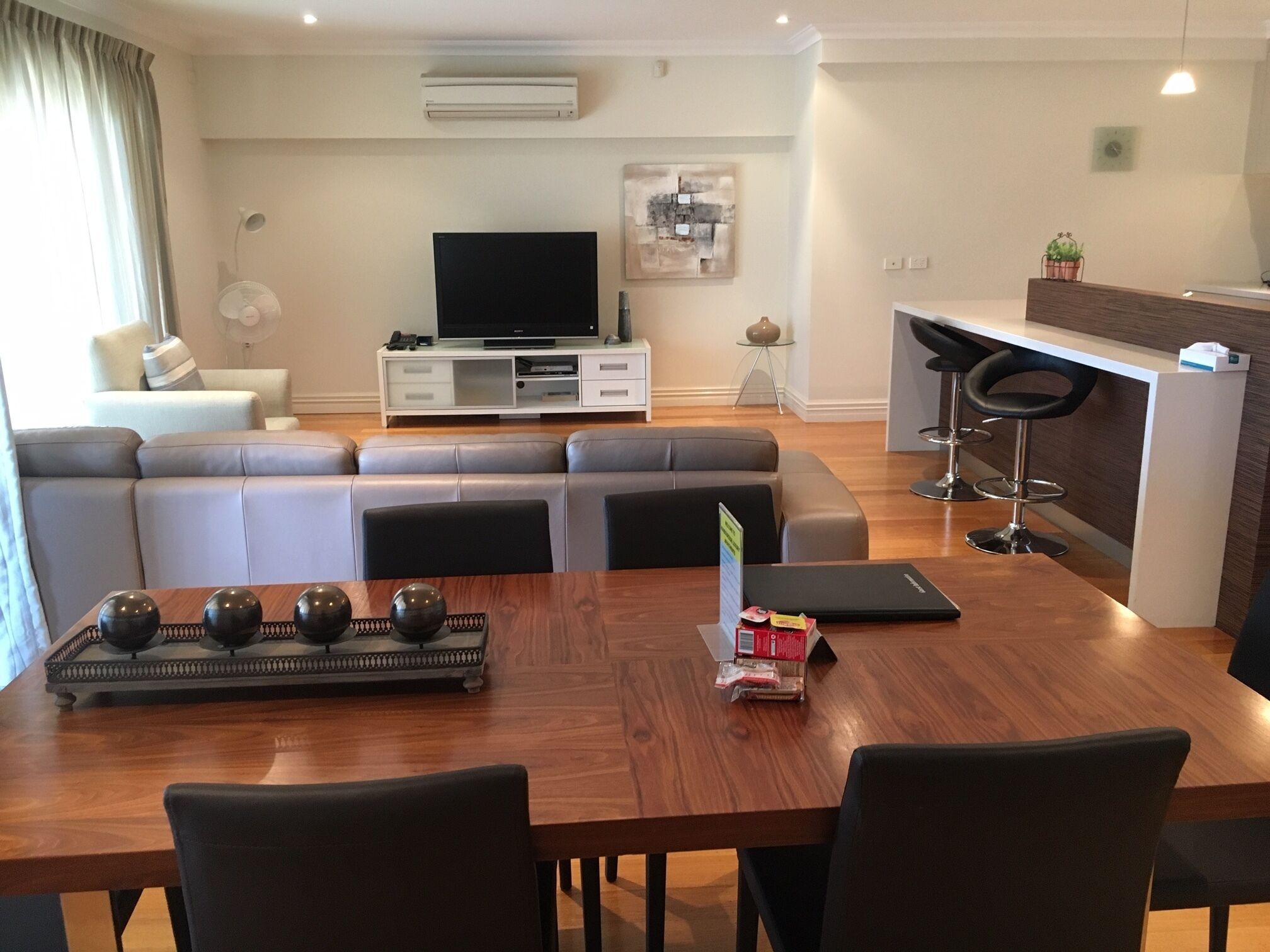 A modern living room with a sofa set, a dining table with chairs, a kitchen area with a bar, and a flat-screen TV on a stand.
