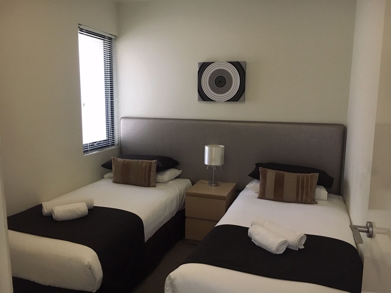 A hotel room with two beds, a bedside table with a lamp, a window with blinds, and a wall-mounted air vent.