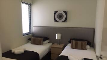 A hotel room with two beds, a bedside table with a lamp, a window with blinds, and a wall-mounted air vent.