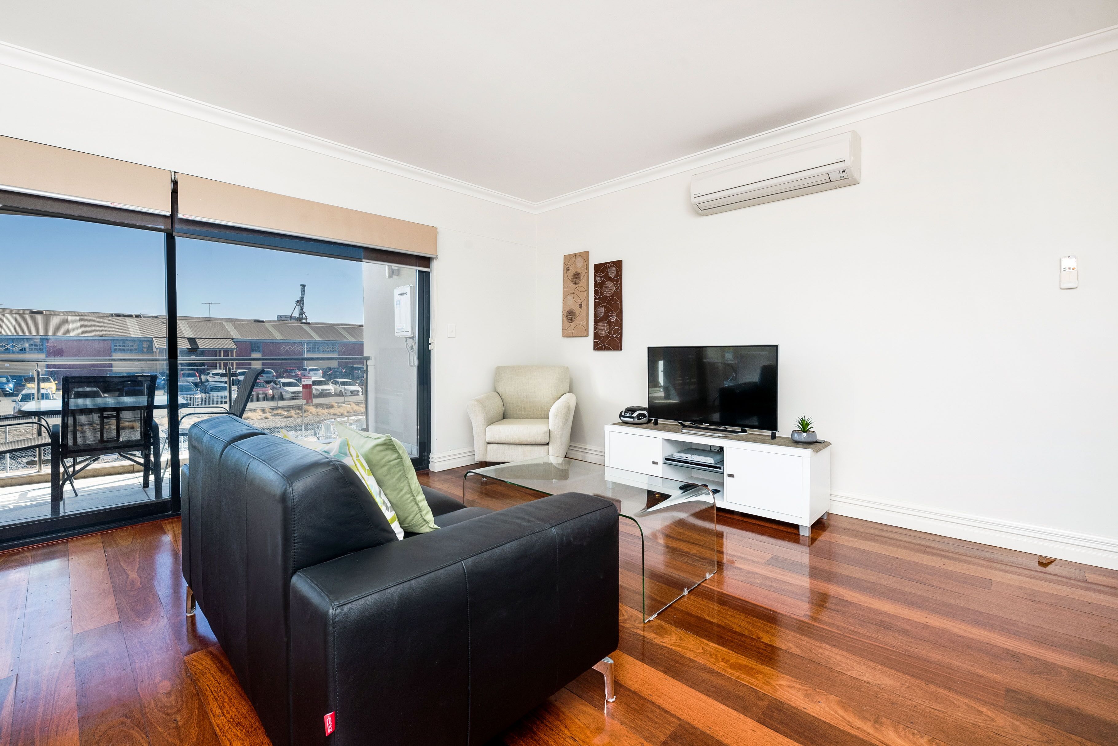 A modern living room with a black leather sofa, a white armchair, a glass coffee table, a flat-screen TV, and a white cabinet.