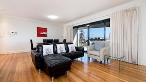 A modern living room with a black leather sofa, a glass coffee table, and a beige armchair. There is a dining area with a table and chairs, and a large window offering a view of the outdoors. - Fremantle Harbourside  Apartments (Fremantle)