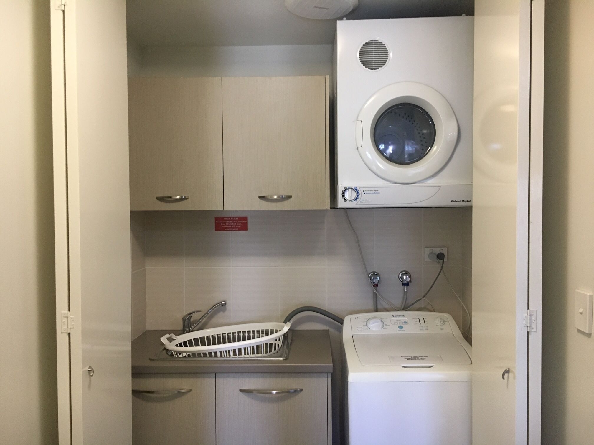 A compact laundry area with a front-loading washer and dryer, a sink with a drying rack, and a cabinet with a 'Do Not Disturb' sign.