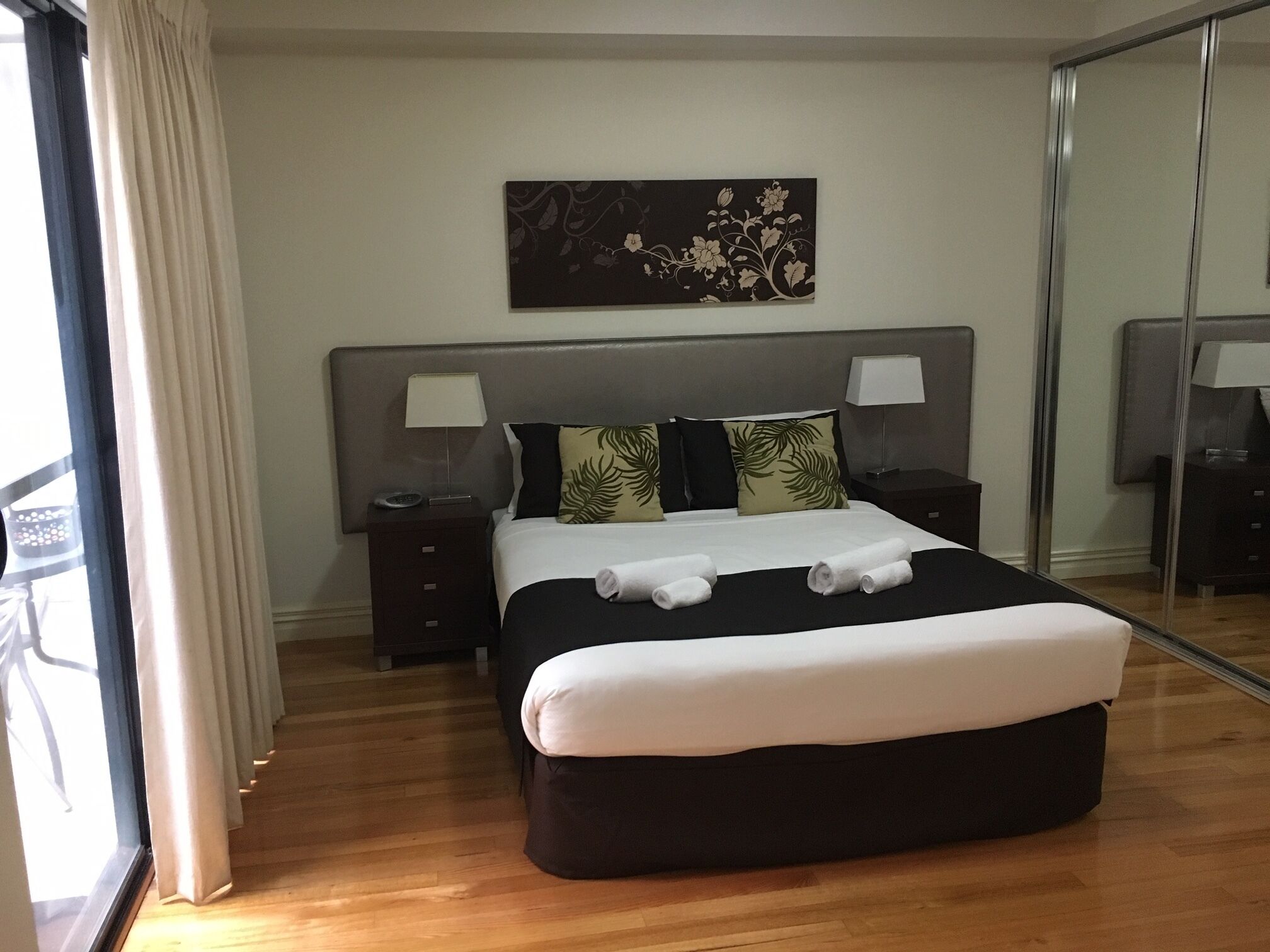 A modern hotel room with a large bed, a nightstand with a lamp, a mirror, and a painting on the wall.