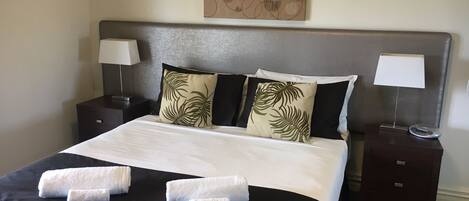 Una cama bien hecha con colcha blanco y negro, dos almohadas con estampado de hojas de palma y dos toallas blancas. Hay dos mesitas de noche con lámparas, una a cada lado de la cama.