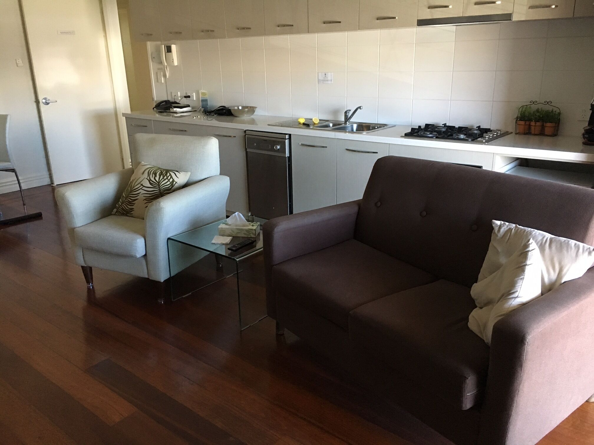 A modern kitchen with white cabinets, a gas stove, and a sink. There is a brown sofa and a white armchair with a glass coffee table in the living area.