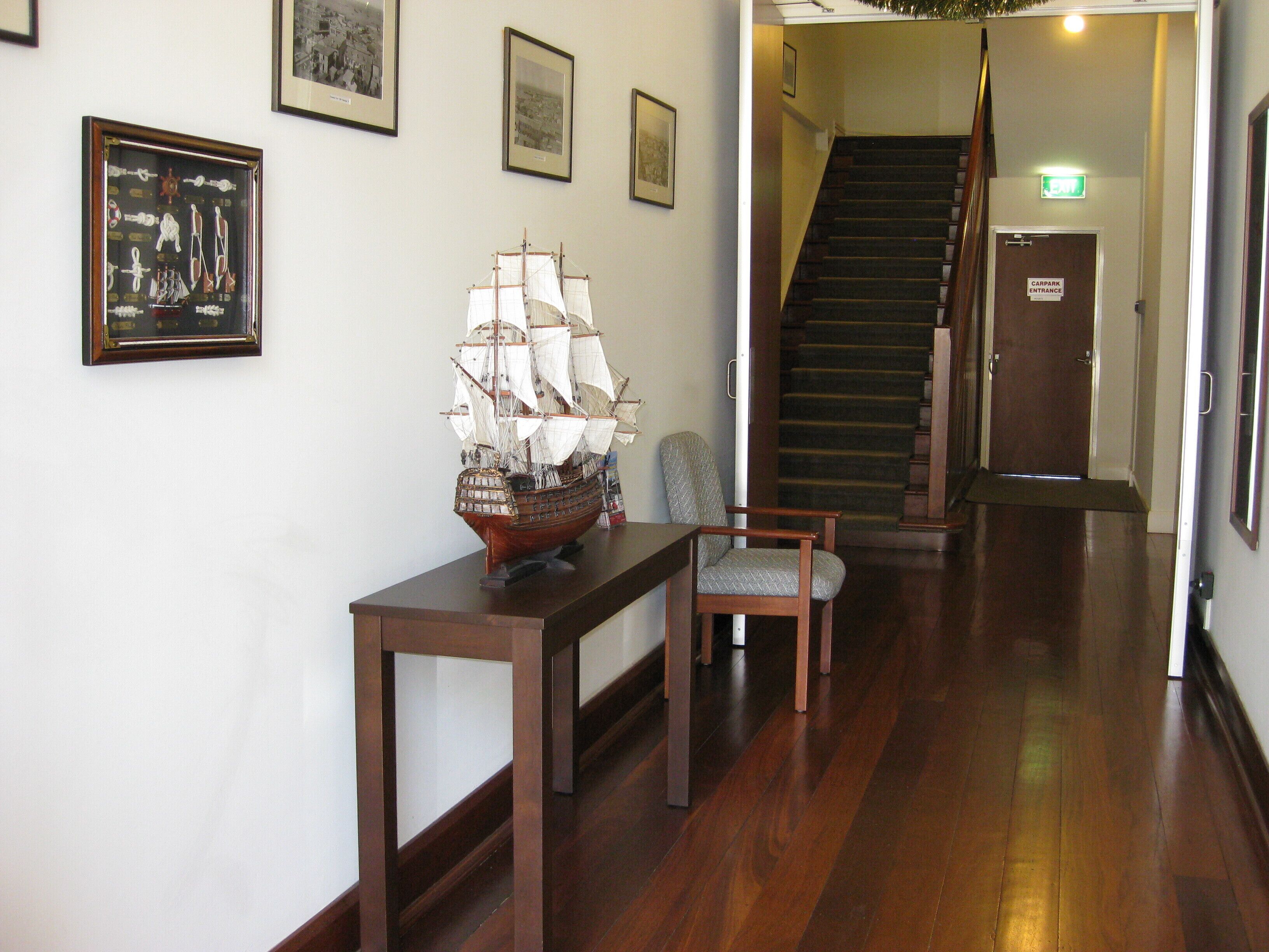 A hallway with a wooden ship model on a table, a chair, and framed pictures on the wall. A staircase is visible in the background.