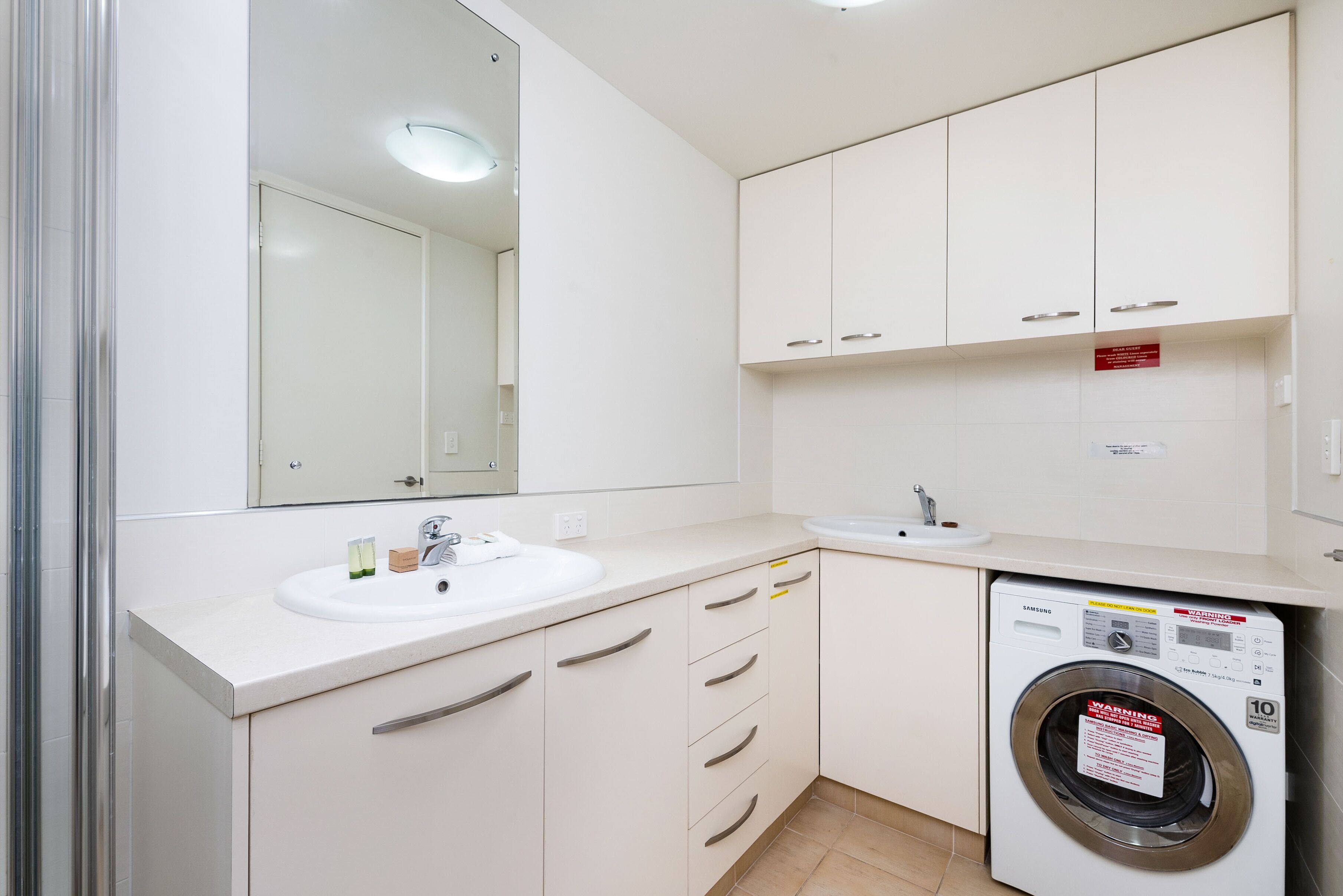 A modern bathroom with a sink, mirror, cabinets, and a front-loading washing machine.