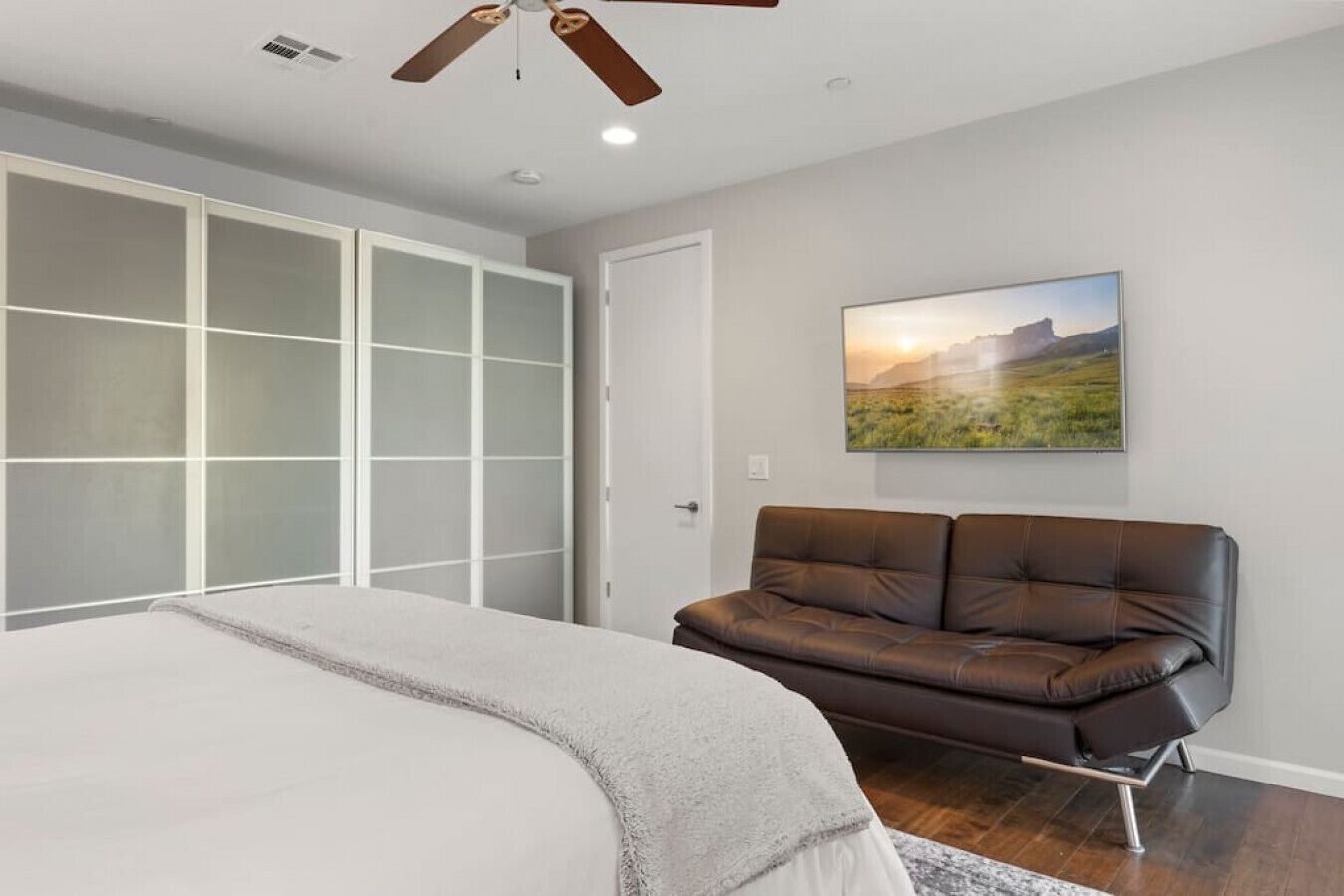 A bedroom with a large bed, a brown leather sofa, a ceiling fan, and a landscape painting on the wall.