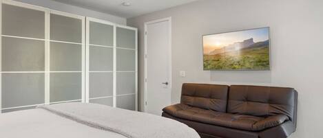 A bedroom with a large bed, a brown leather sofa, a ceiling fan, and a landscape painting on the wall.