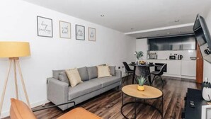 Ein modernes Wohnzimmer mit einer grauen Couch, einem runden Couchtisch und einer Stehlampe. Es gibt eine Küchenzeile mit einem Esstisch und Stühlen.