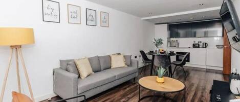 Una sala de estar moderna con un sofá gris, una mesa redonda de centro y una lámpara de pie. Se observa una zona de cocina con una mesa de comedor y sillas.