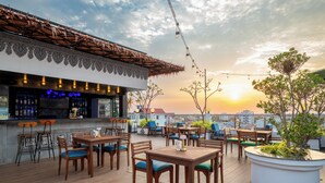Un bar sul tetto con tavoli e sedie in legno, un bancone bar con bottiglie e vista sul tramonto.
