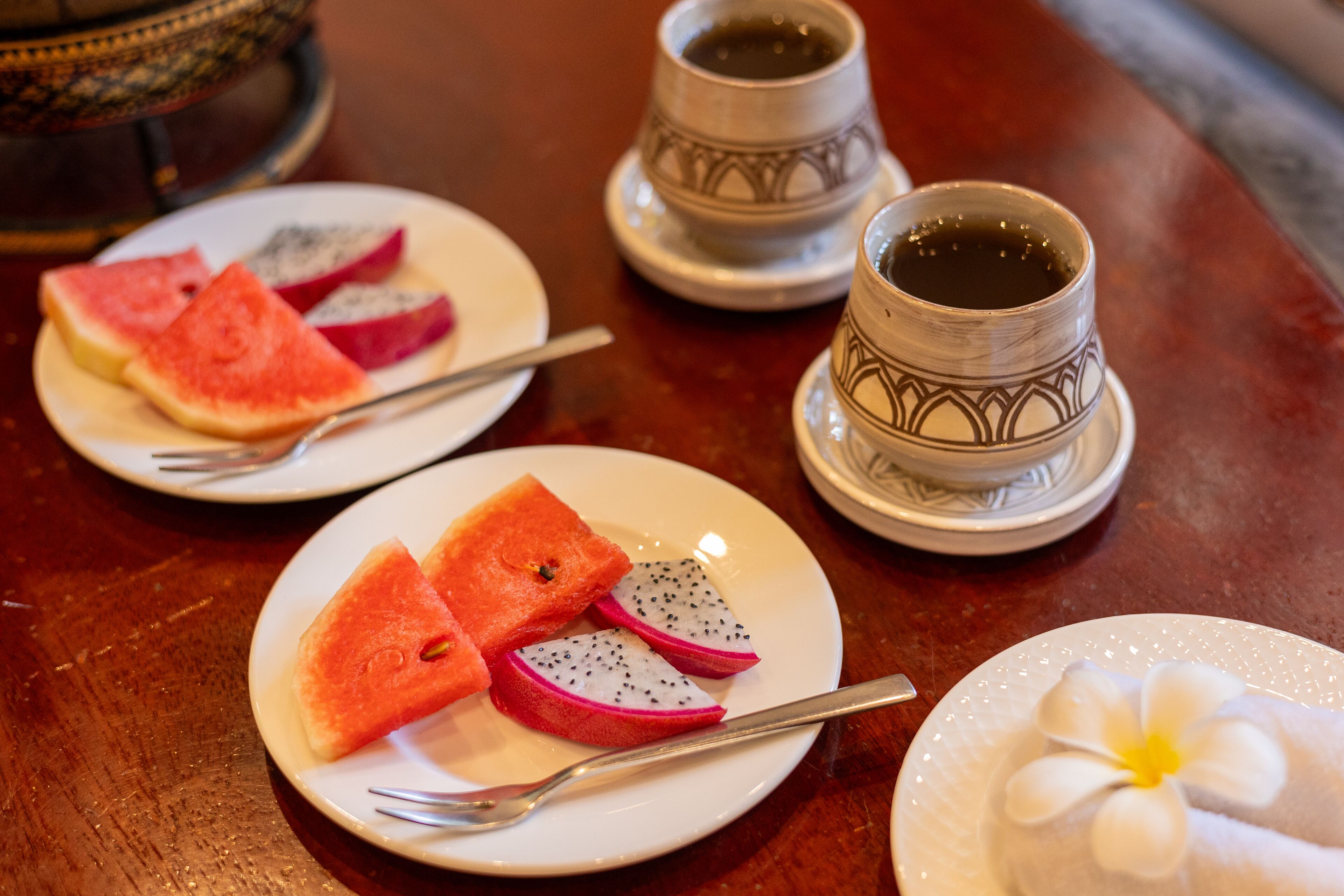 A table with plates of watermelon and dragon fruit, a cup of tea, and a decorative flower.