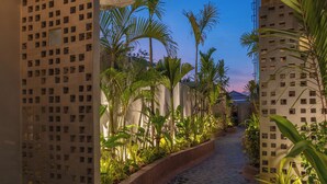 Un sentiero stretto, delimitato da rigogliosa vegetazione e edifici con facciata a griglia.
