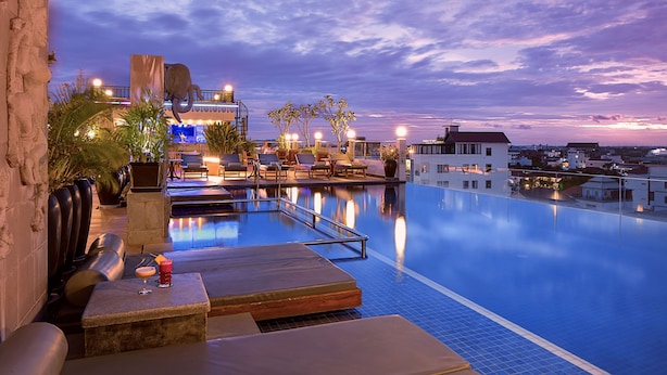 Una piscina a sfioro con poltrone relax e un'area bar, circondata da edifici e un cielo al tramonto.