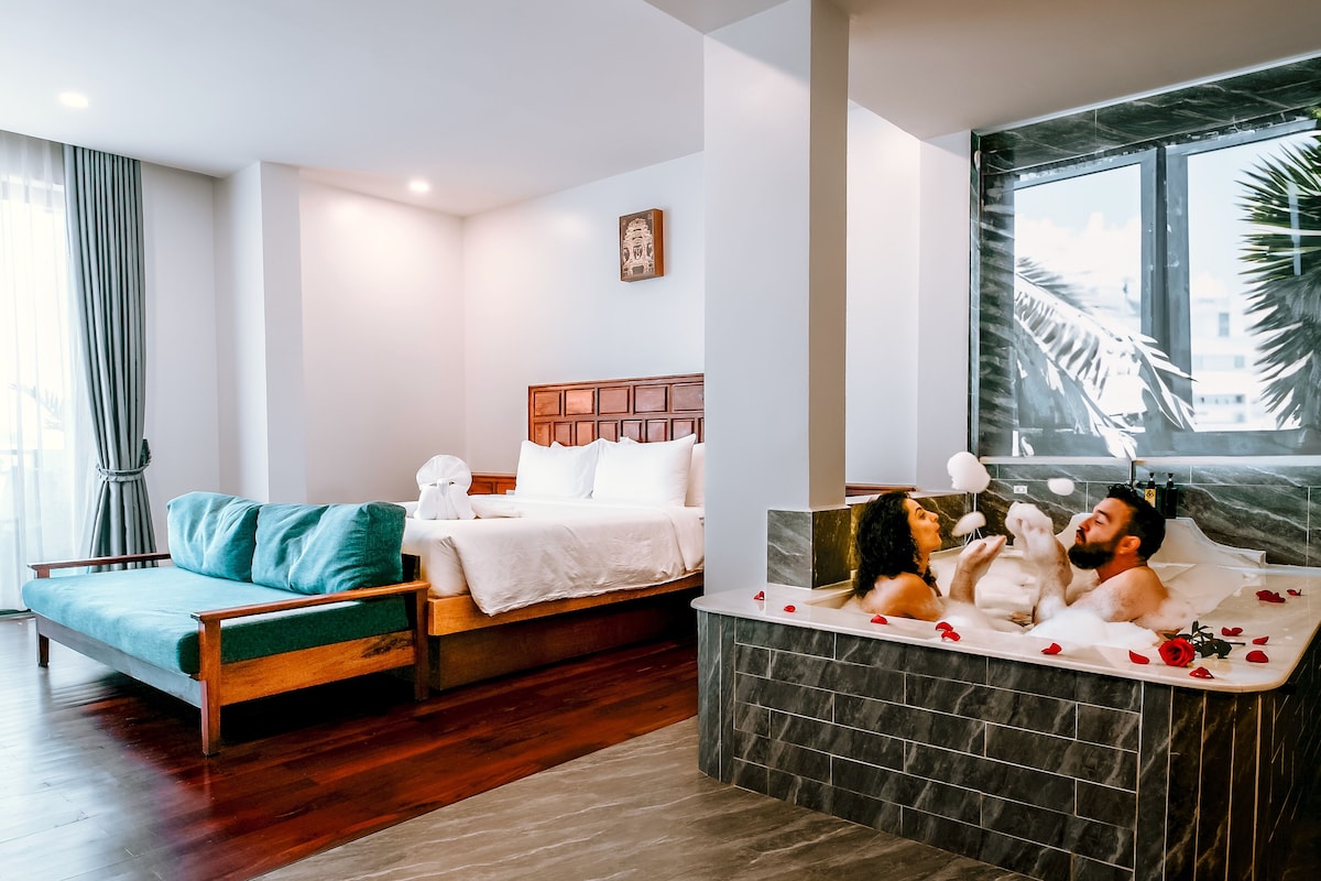 Hotelkamer met een bed, een slaapbank, een badkuip met rozenblaadjes en een groot raam met uitzicht op palmbomen.