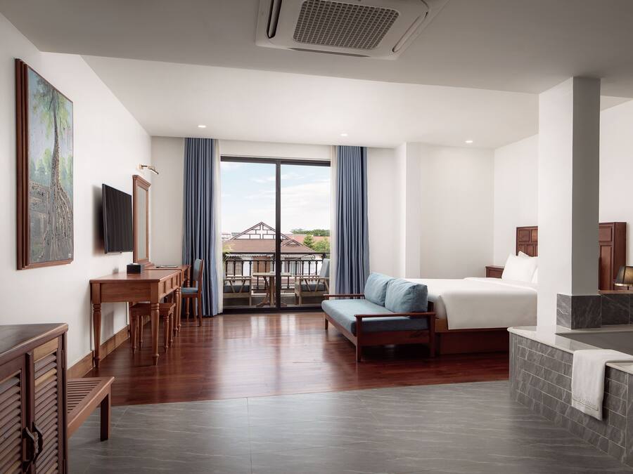 Una habitación de hotel moderna con una cama grande, un área de estar con un sofá y sillones, una mesa de comedor, un televisor y vistas al exterior.