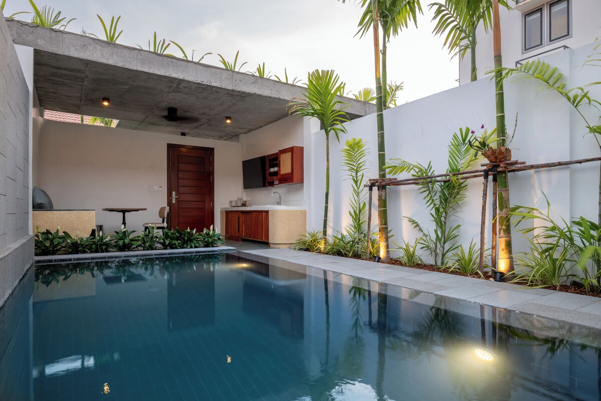 A modern outdoor pool area with a concrete structure, a wooden door, and a kitchen area with cabinets and a TV.
