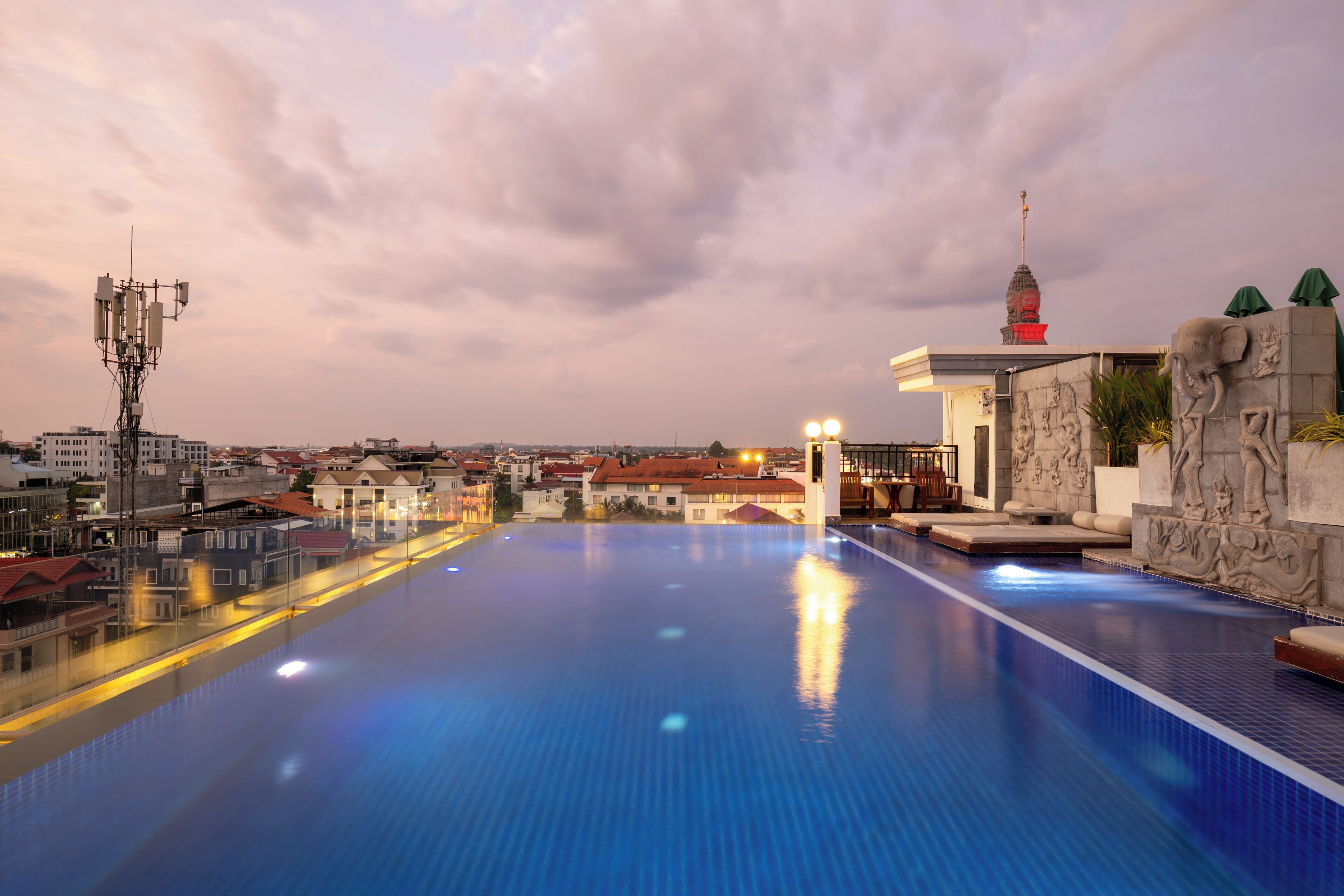 A rooftop infinity pool with a city view, a stone wall with elephant sculptures, and a building with a red roof.