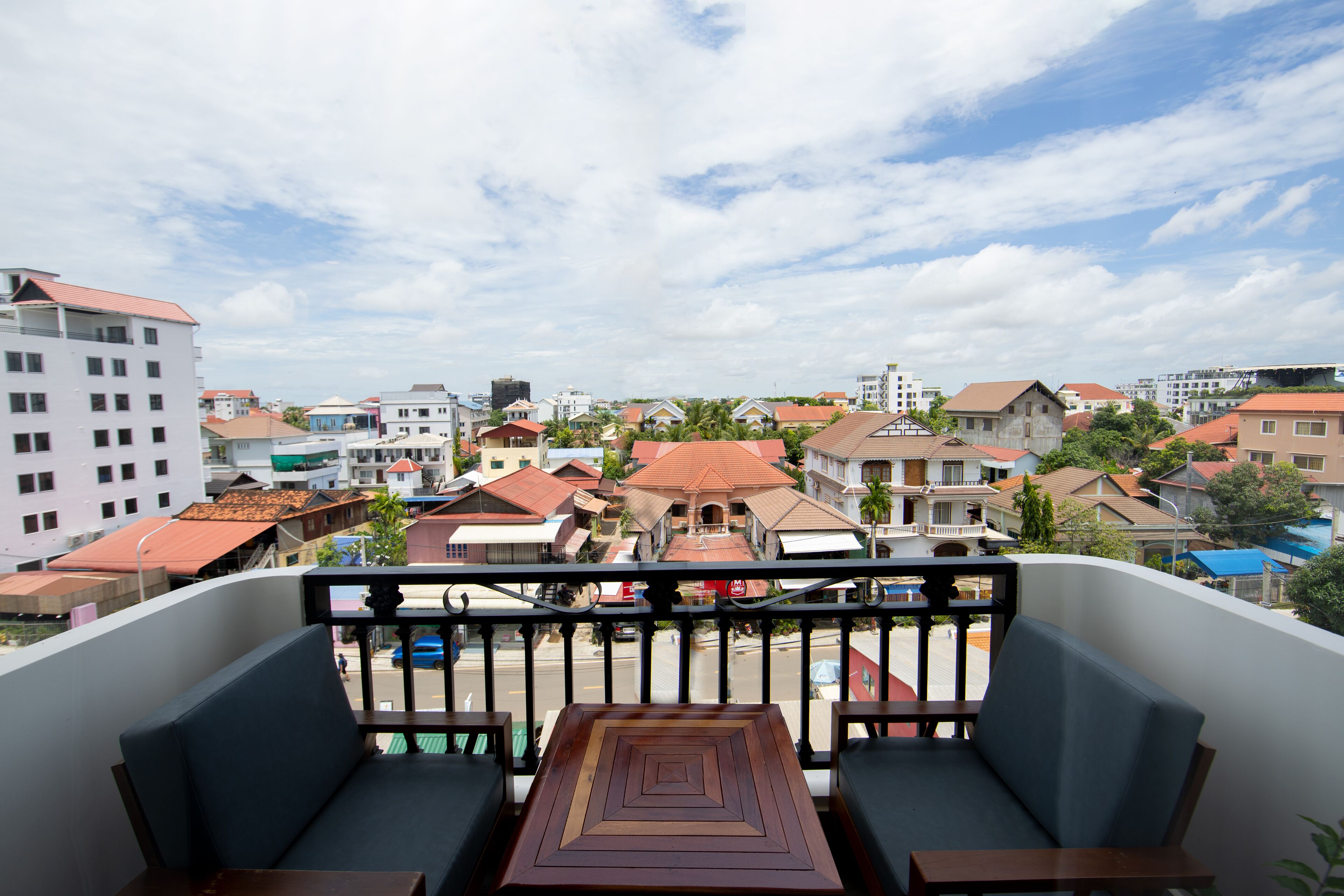 A balcony with a wooden table and two chairs, overlooking a residential area with various buildings and a clear sky.