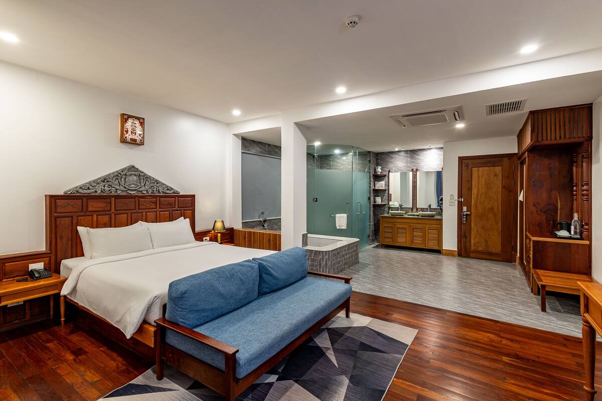 Une chambre d’hôtel moderne avec un lit en bois, un canapé bleu, une salle de bain avec baignoire et une armoire en bois.