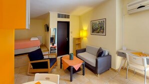 Un espacio habitable pequeño y funcional con cama, sofá, mesa y escritorio. Hay una lámpara y un cuadro en la pared.