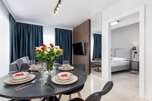 A modern dining area with a round table set for four, a vase with flowers, and a TV in the background.