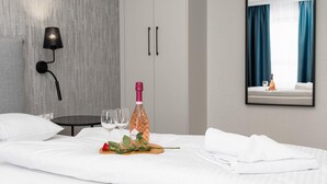 Habitación de hotel con una cama, una mesita de noche con dos vasos, una botella de champán y un espejo que refleja una ventana con cortinas.