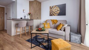 A modern living room with a sofa, two bar stools, a coffee table, and a small ottoman. A kitchen area is visible in the background with a wooden cabinet and a hanging plant. - Modern Studio in Mielno | View of the Sea and Parking Lot (Mielno)