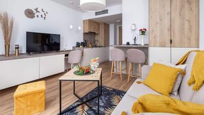 A modern living room with a sofa, coffee table, and a TV. There is a kitchen area with wooden cabinets and a dining space with chairs. - Modern Studio in Mielno | View of the Sea and Parking Lot (Mielno)