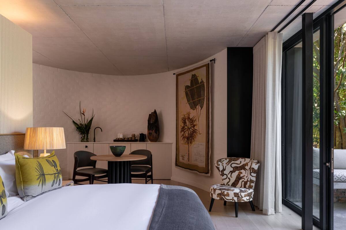 Camera d'hotel moderna con un letto, un tavolo da pranzo, sedie, una poltrona con motivo decorativo e un'ampia finestra che dà su un panorama esterno.