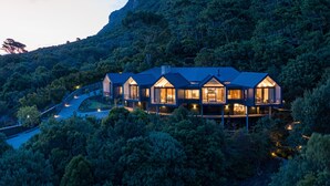 Una casa moderna con varias habitaciones, rodeada por un bosque frondoso y una montaña al fondo.
