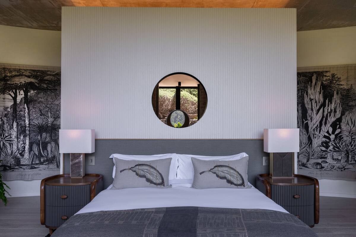 Una camera da letto con un letto grande, due comodini, uno specchio rotondo e un’opera d’arte a parete raffigurante una scena di bosco.