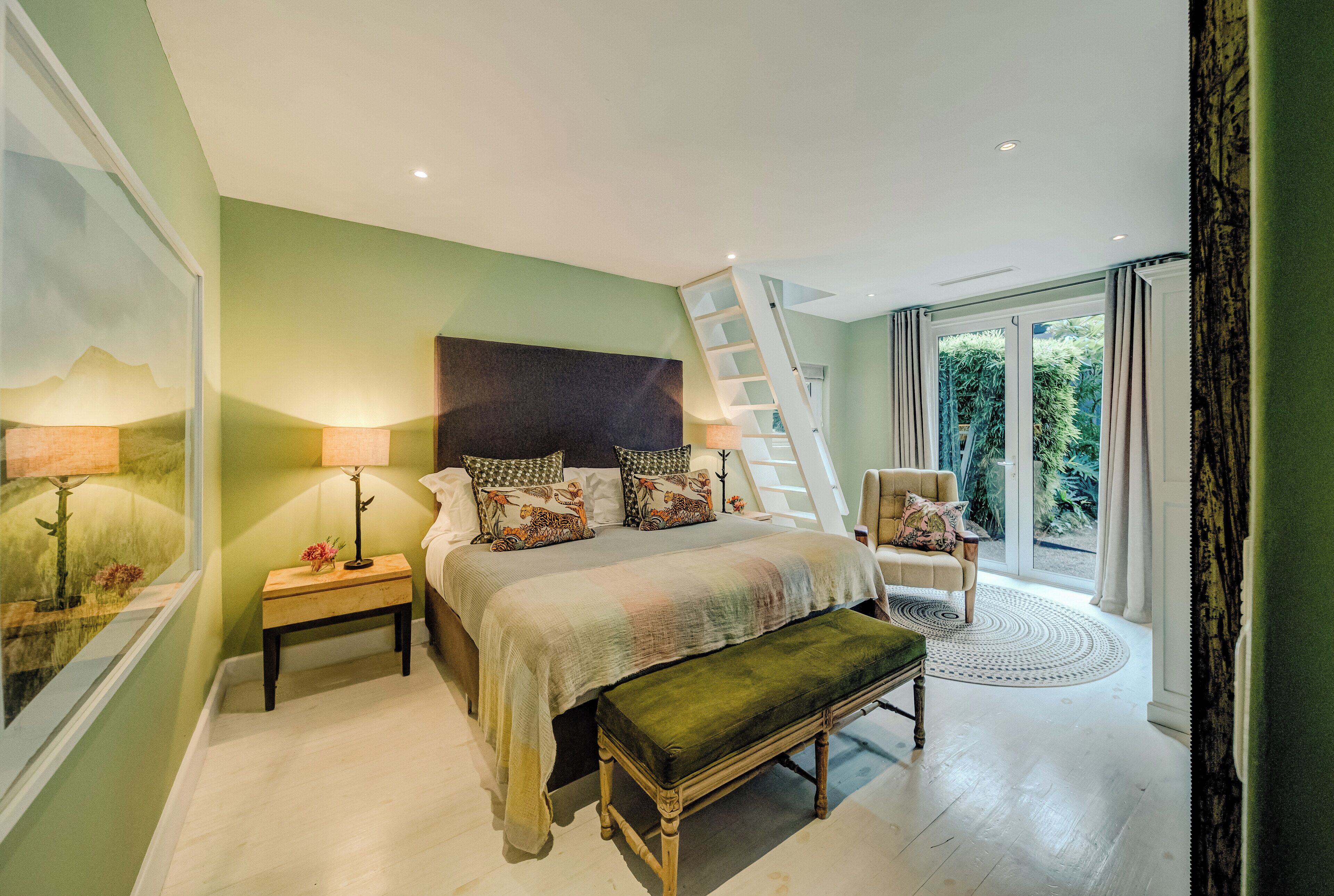 A bedroom with a bed, a bench, a nightstand, a chair, a ladder, and a view of greenery outside.