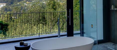 Un baño moderno con una bañera independiente, una mesita pequeña y una puerta de vidrio que da a un balcón con vistas a un paisaje montañoso.