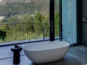 A modern bathroom with a freestanding bathtub, a small table, and a glass door leading to a balcony with a view of a mountainous landscape.
