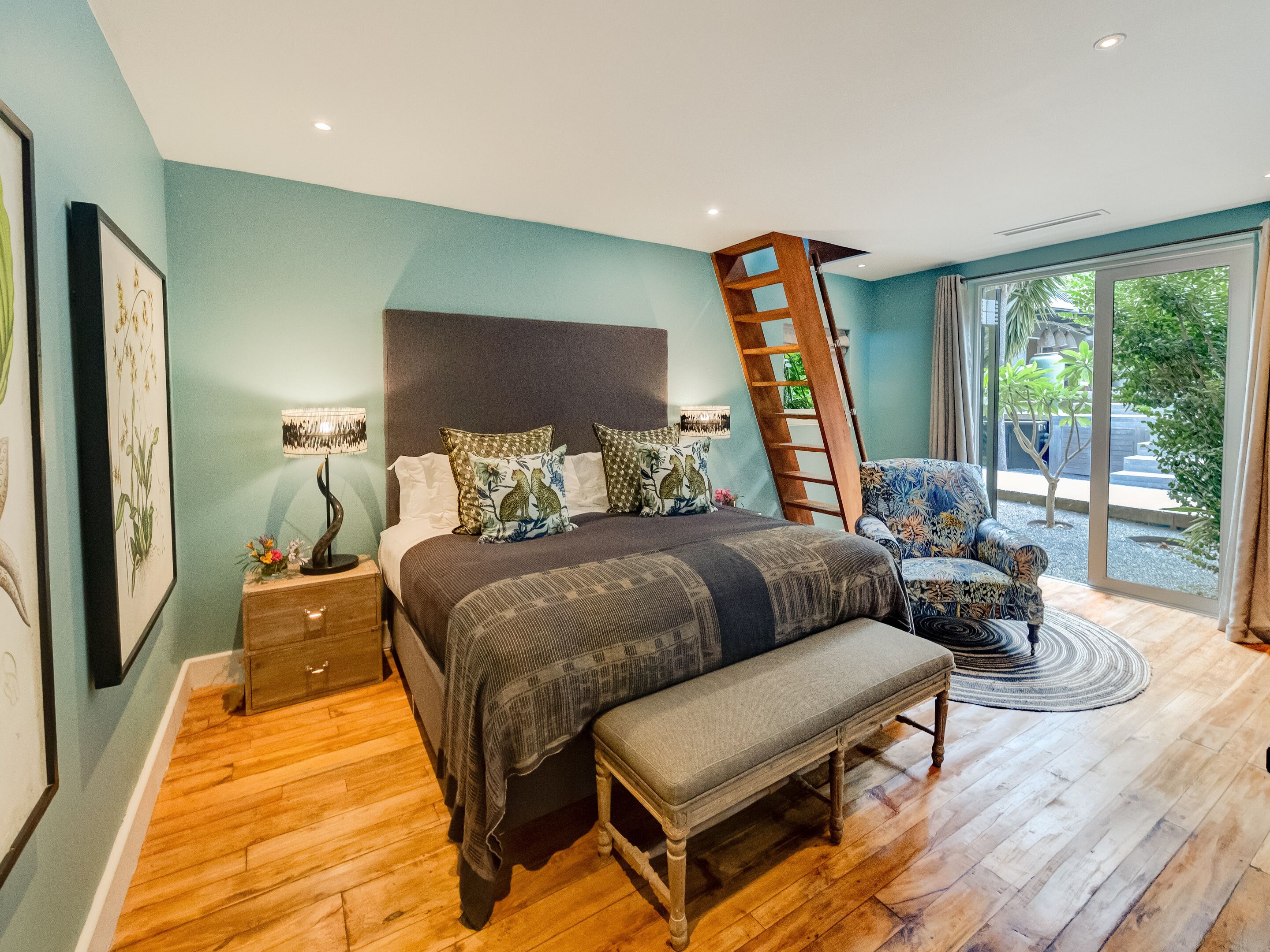 A bedroom with a bed, bedside tables, a bench, a chair, a ladder, and a view of the outdoors through a large window.
