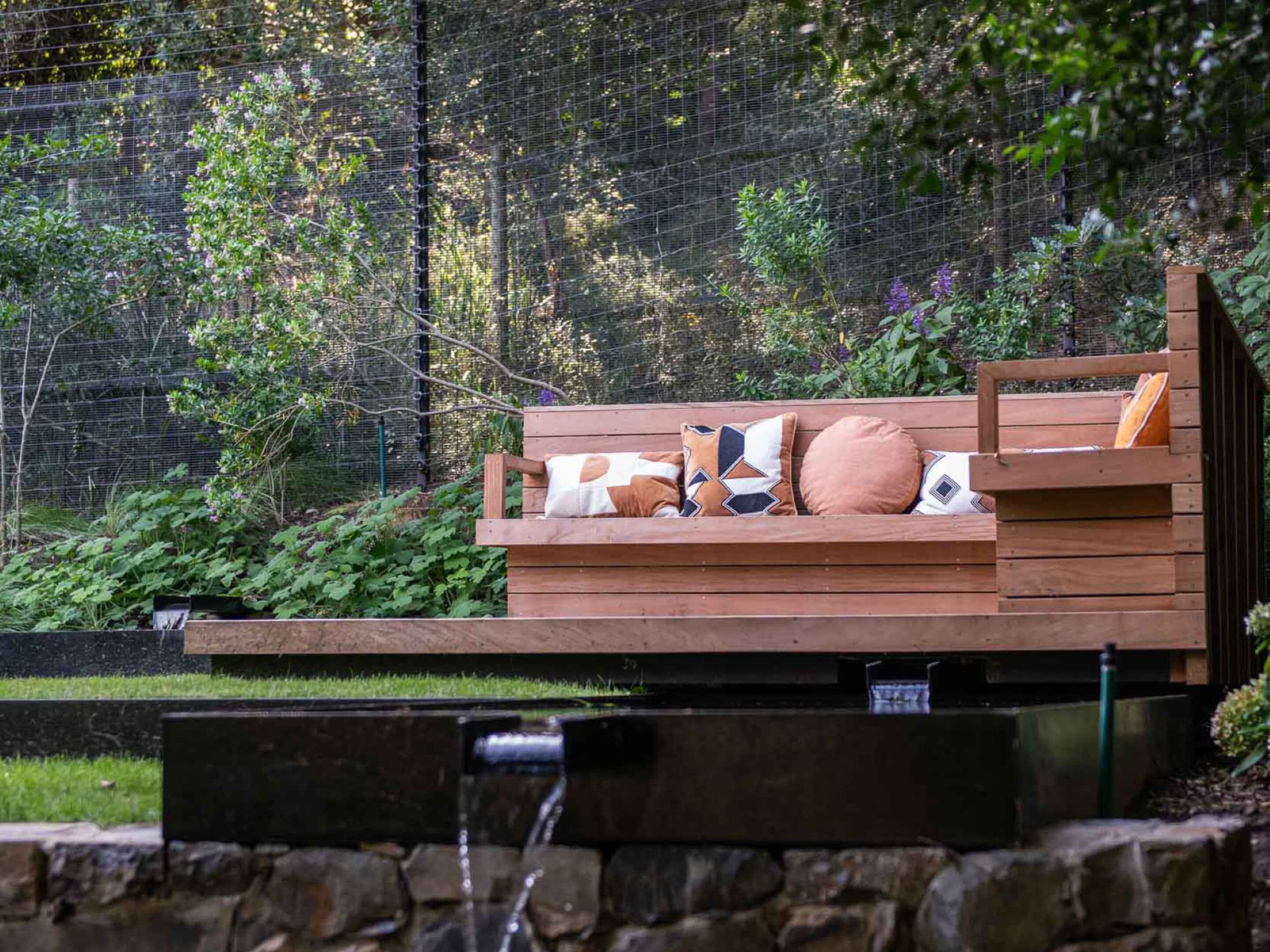 A wooden bench with cushions, surrounded by greenery and a wire fence.
