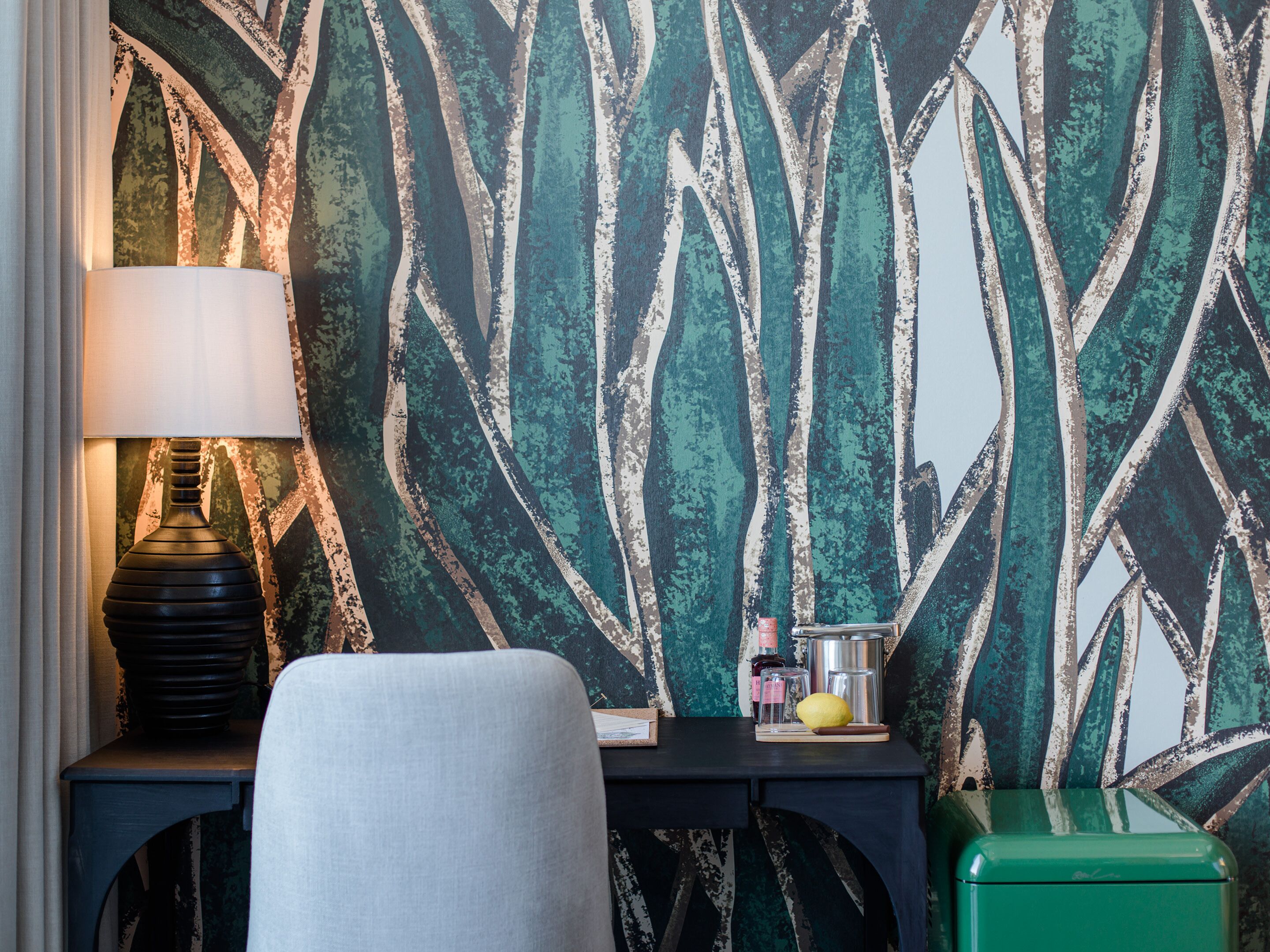 A room with a patterned wallpaper, a lamp, a chair, a small table with a lemon, and a green suitcase.