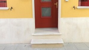 A red door with a small porch, a wall-mounted light fixture above it, and a window with shutters on the side. - Appartamento Nuovo A Chioggia Vista Laguna (Chioggia)