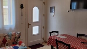 A room with a sofa, a dining table with chairs, a door with a window, and a wall-mounted light fixture. - Appartamento Nuovo A Chioggia Vista Laguna (Chioggia)