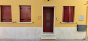 A building with a yellow facade, a red door, and two windows with red shutters.