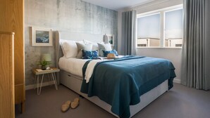 Een slaapkamer met een bed, nachtkastje, lamp, lijst met afbeelding en een raam met gordijnen.