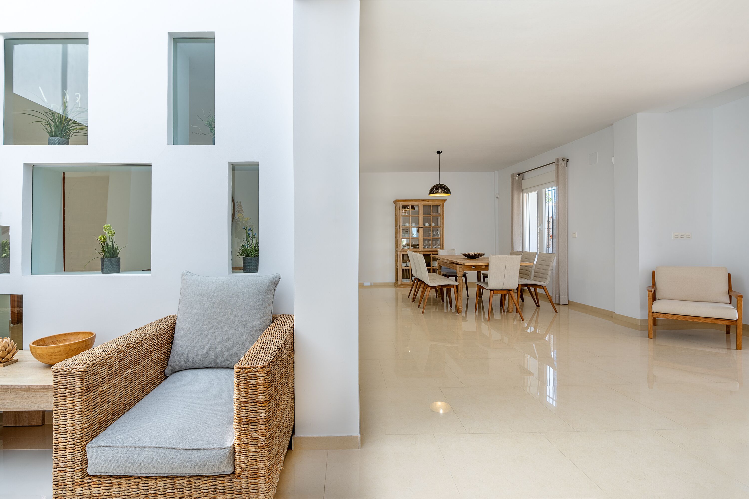 Un soggiorno moderno con un divano in vimini, un'area pranzo con un tavolo e sedie in legno, e un'ampia finestra con piante.