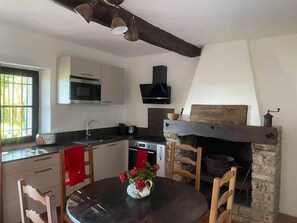 A kitchen with a stone fireplace, wooden chairs, a dining table with a vase of flowers, and a modern microwave.