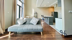 A modern living room with a grey sofa, two decorative pillows, a dining area with a table and chairs, and a kitchen area with a refrigerator and bar stools. - Homesuite’ Home @ Riverson SOHO (Kota Kinabalu)